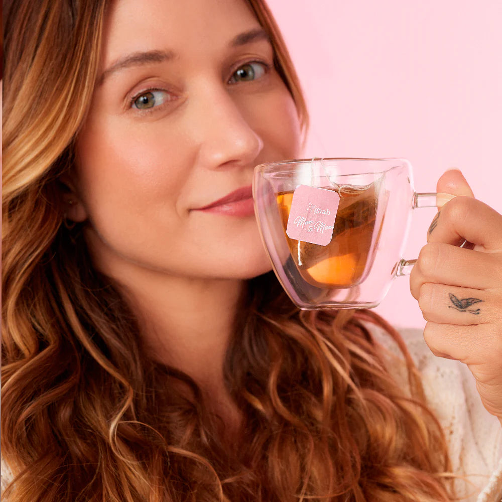A woman with a gentle smile holds a clear mug of GAAB Mom to Mom tea, emphasizing a moment of tranquility and self-care.