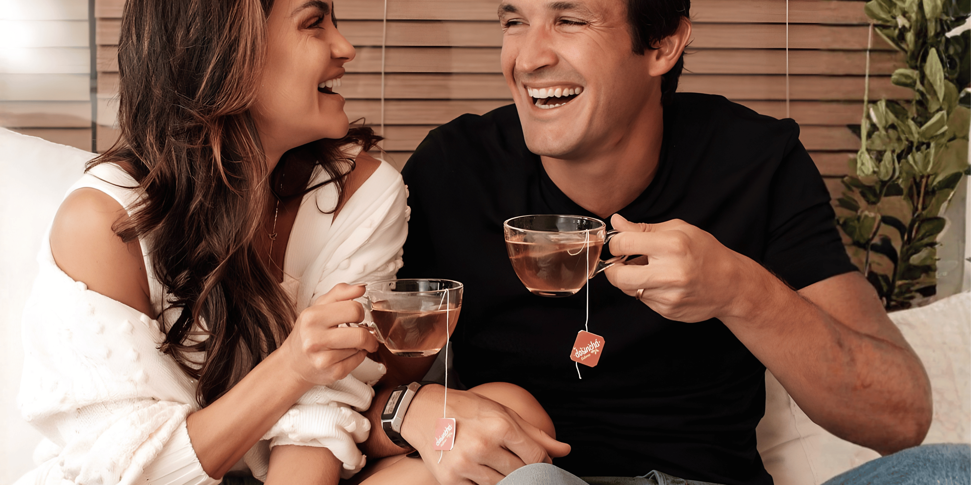 A smiling couple sitting closely together on a couch, each holding a glass mug with a desincha tea bag hanging from it. The woman wears a white off-the-shoulder top, and the man wears a black T-shirt.