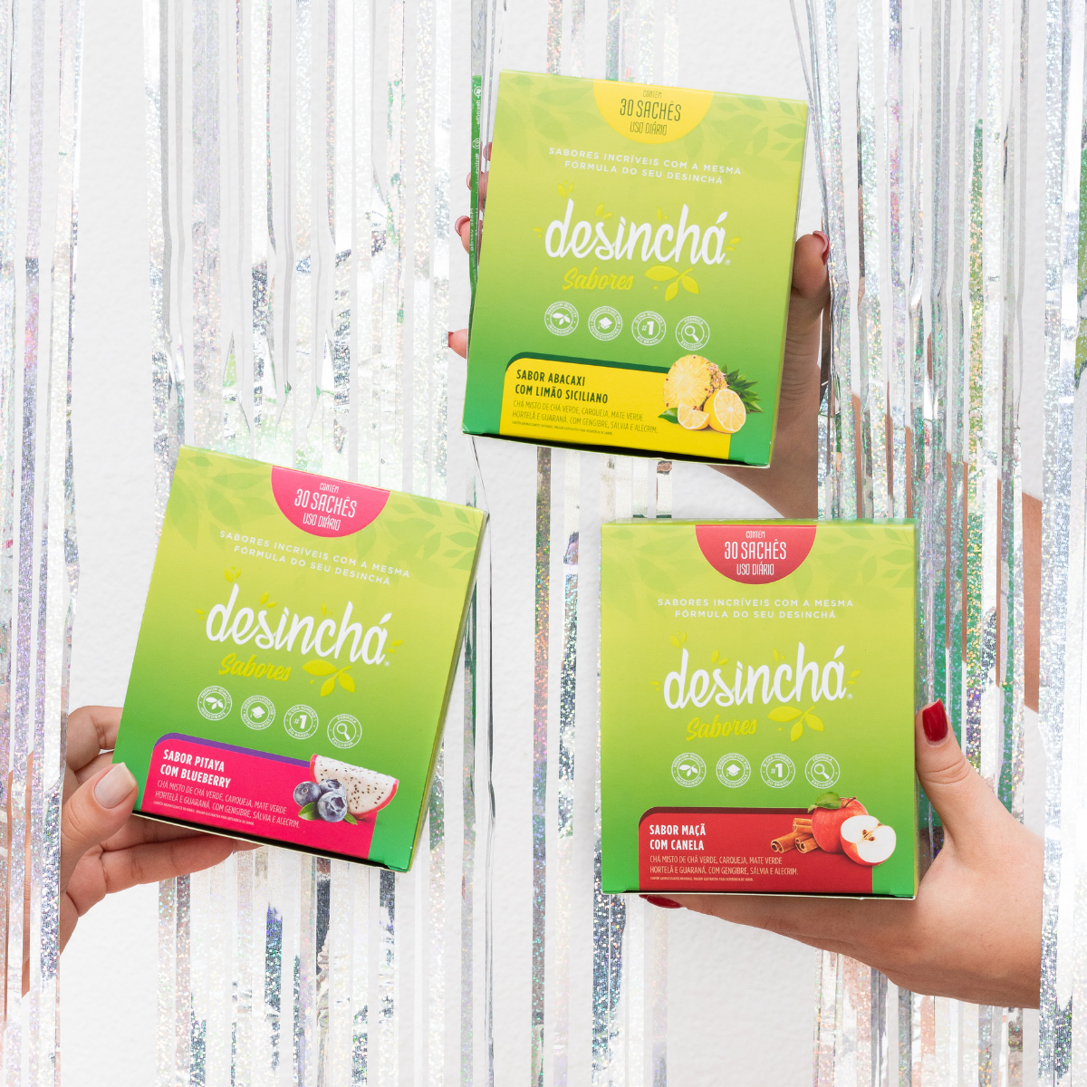 Three hands holding boxes of desinchá teas against a white tinsel background. The boxes are green and yellow with pink and red accents, showing different flavors.