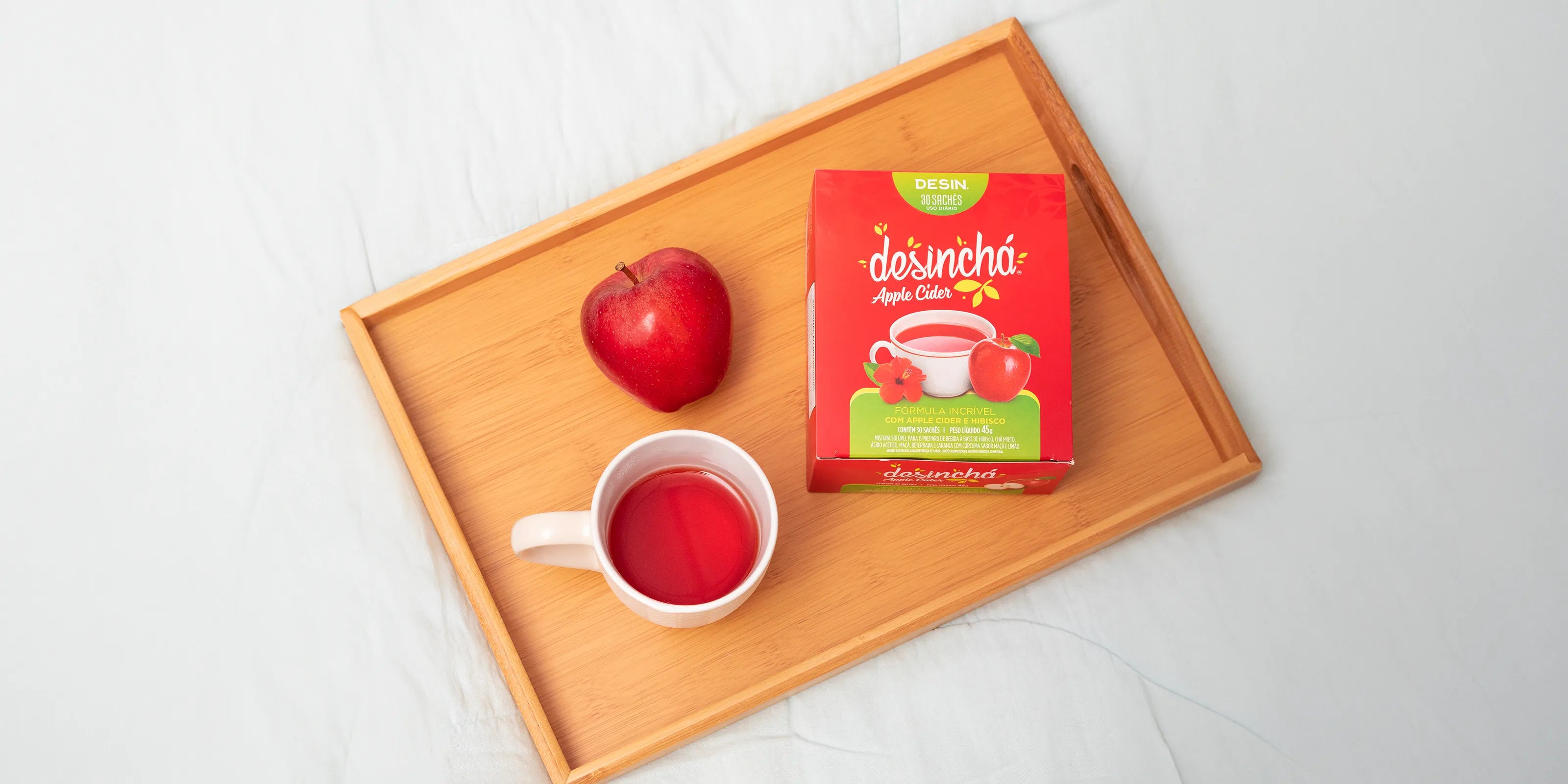 Desinchá Apple Cider tea with a red apple and a mug of brewed tea on a wooden tray against a white background.