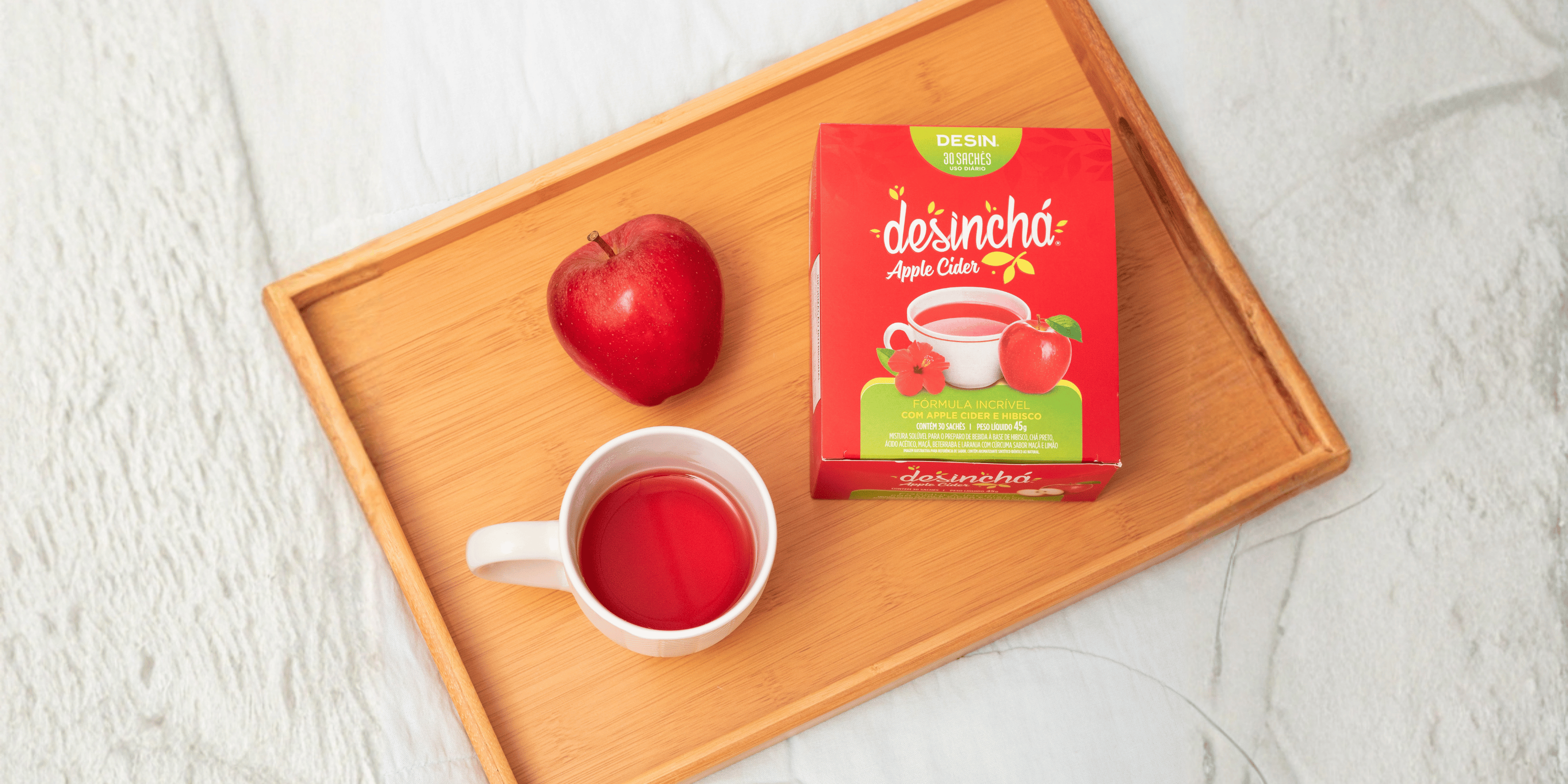 A red box of desincha Apple Cider tea sits on a light wood tray next to a white mug filled with red tea and a red apple, all on a light gray marble surface.
