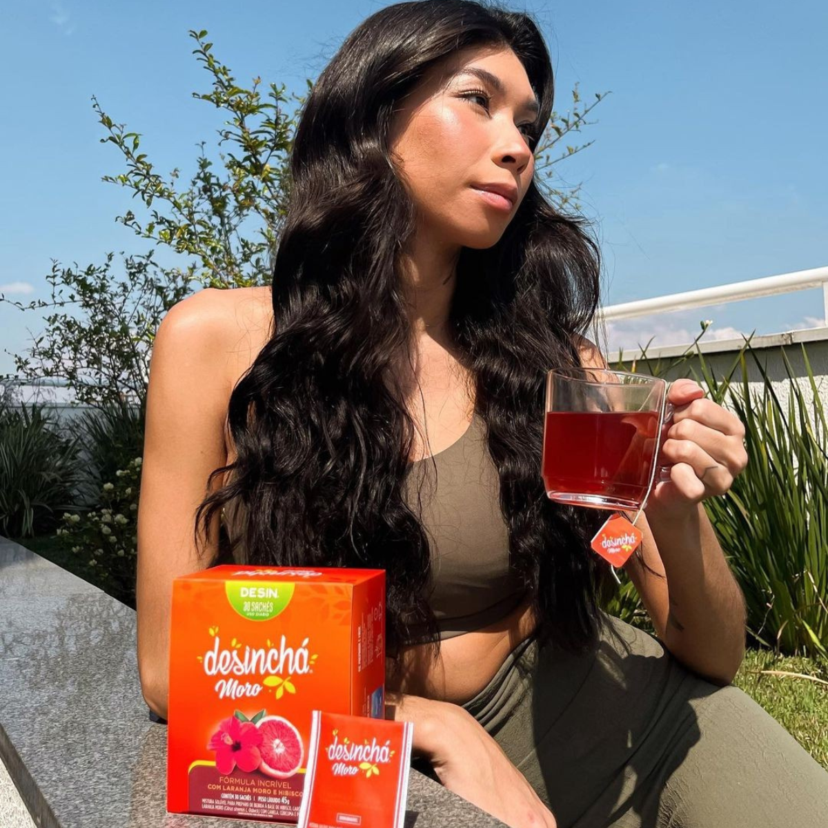 A woman outdoors holding a glass of red tea next to a box of desincha orange and hibiscus tea bags, wearing an olive green sports bra and matching pants under a blue sky.