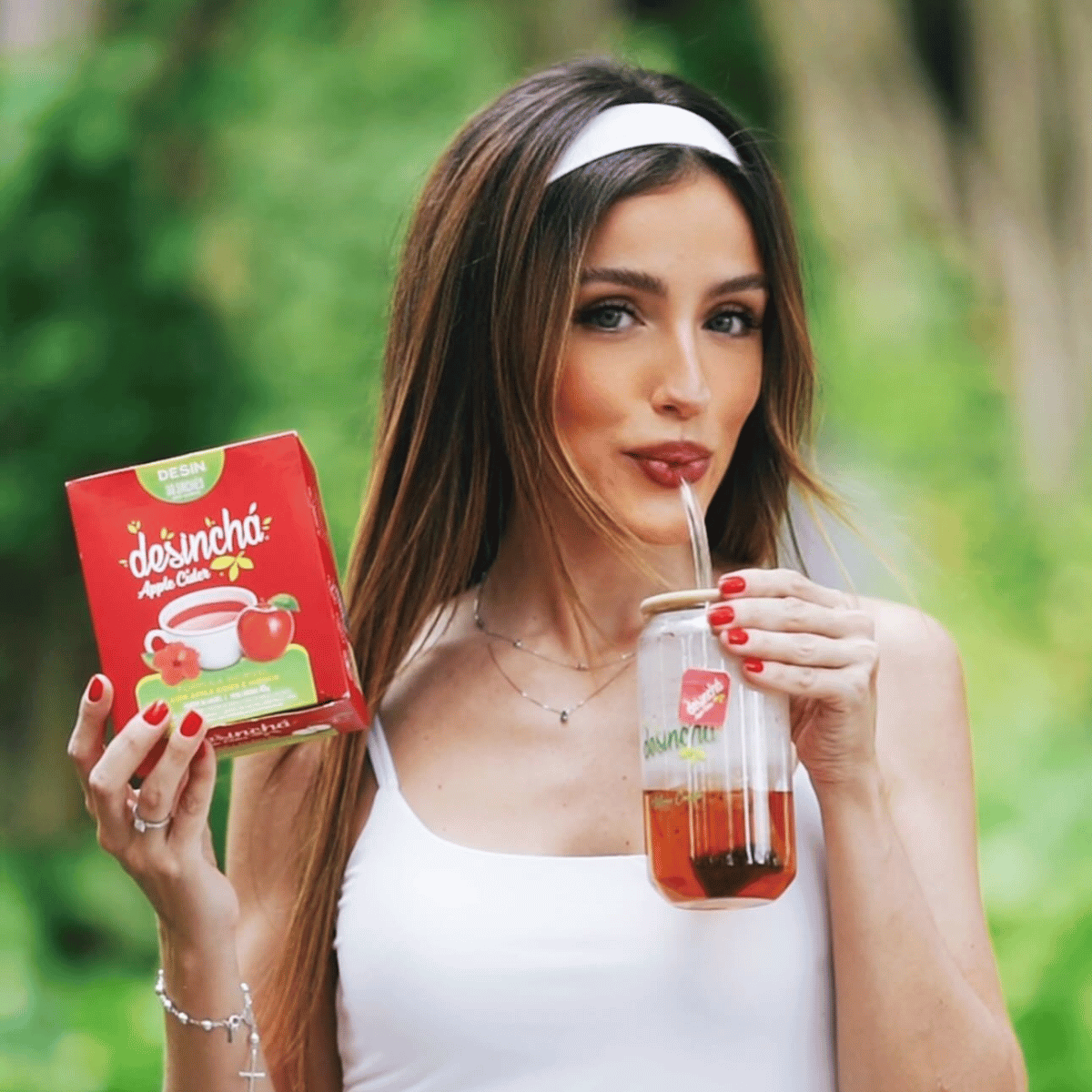 A woman outdoors holds a red box of desincha's apple tea and a glass jar of the tea with a straw. The background is a soft, blurred green garden.