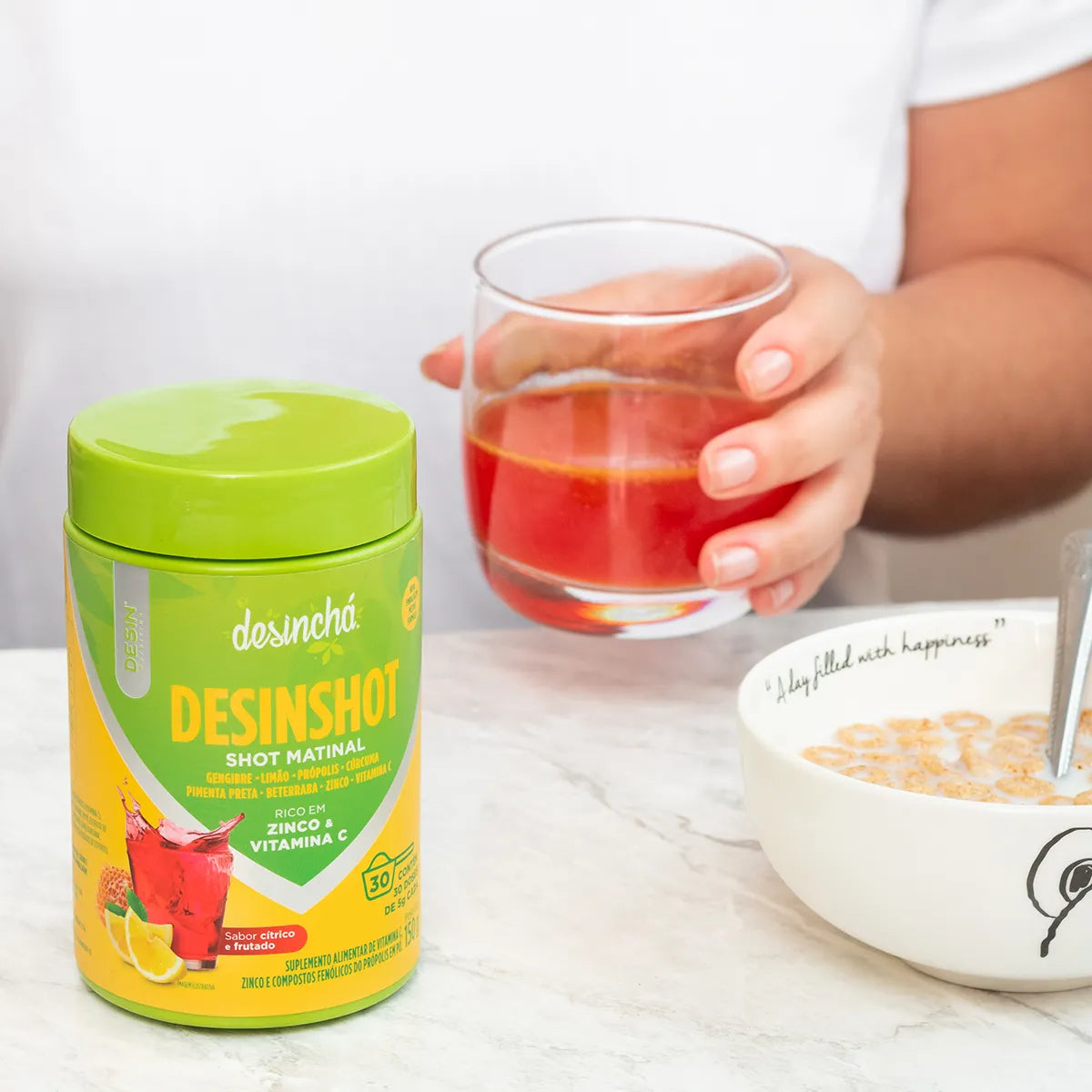 Close-up of a person's hands holding a glass of bright red Desinchá Desinshot next to the product container, suggesting a healthy and energizing morning routine.