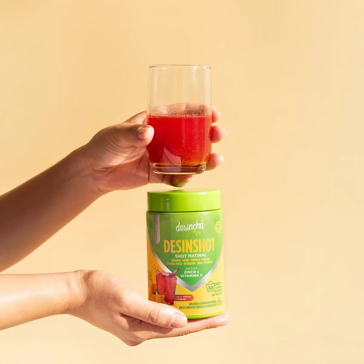 Hand holding a glass of fizzy red morning shot above a container of Desinchá Desinshot, representing a healthy start to the day with a citrusy beverage.