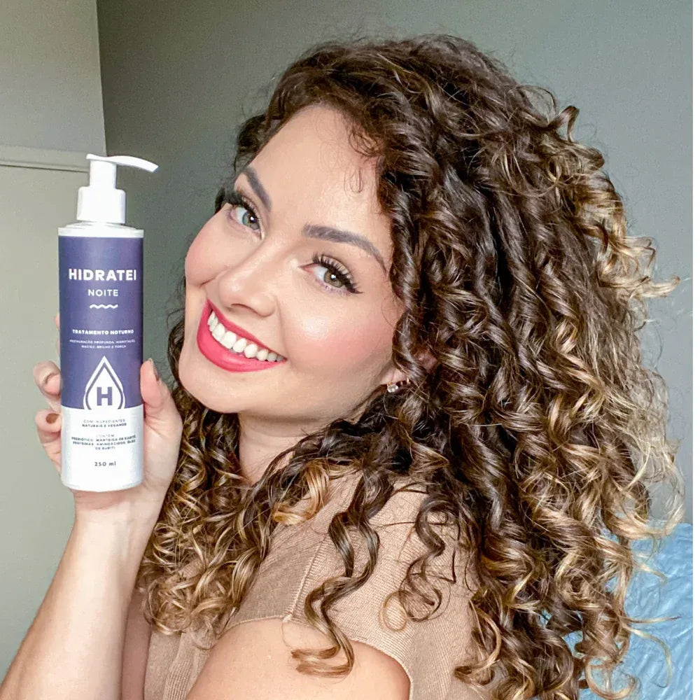 A smiling woman with curly hair proudly showcasing a bottle of Hidratei Night hair treatment, radiating confidence in her beauty regimen.