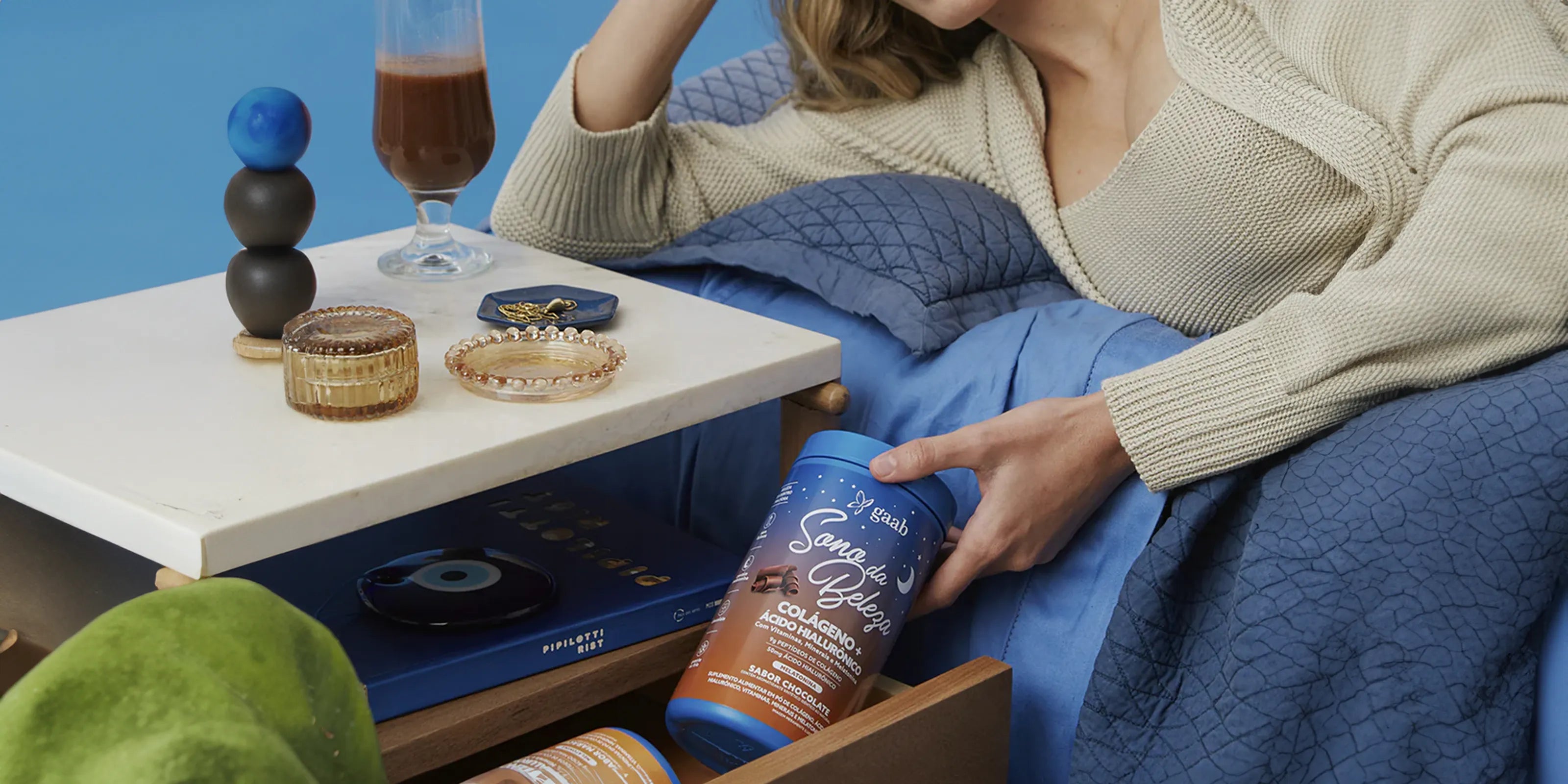 Cozy lifestyle scene with a person relaxing, holding a container of GAAB Sleep Beauty Collagen+ supplement, accompanied by a glass of chocolate-flavored drink and stylish home decor on a side table.